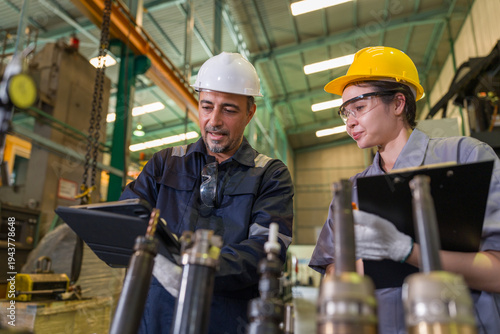 Two diverse engineers wearing hard hats looking at digital tablet discussing project plans in heavy industrial manufacturing factory plant workshop. Professional teamwork safety first concept