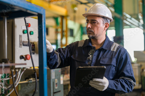 Male industrial engineer holding digital tablet while operating control panel switch in manufacturing factory plant. Professional technician checking machinery system maintenance process