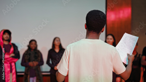 African young adult gen z male man hold scripts paper read play story lines scene standing indoor stage hall. Happy group boy girl do work drama practice day studio campus place