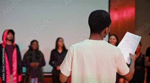 African young adult gen z male man hold scripts paper read play story lines scene standing indoor stage hall. Happy group boy girl do work drama practice day studio campus place