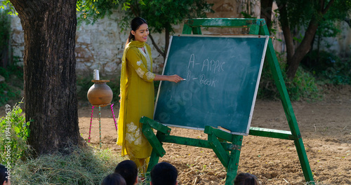 Indian young adult gen z female lady talk speak teach to villager child day under tree rural area. Girl boy group sitting on mat do study look board learn read english at open class field place