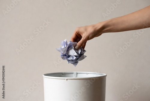 A hand is discarding a crumpled paper ball into a trash can. A simple, neutral background setting.