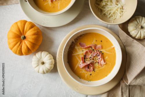 Close-up of a cozy autumn dinner setting featuring homemade butternut squash soup on a linen tablecloth with small white and orange pumpkins and shredded cheese in a ceramic bowl.