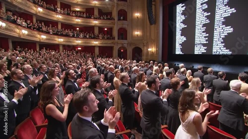 Audience applauds enthusiastically at a formal event in a grand theater with ornate balconies and a large screen displaying text.