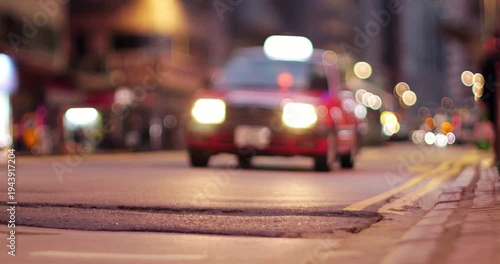 Red taxi ride towards camera at night street of Hong Kong Island, tilt-shift shot with blurred top of frame. Scenic gently blinking lights in bokeh on background, typical scenery of Western district.