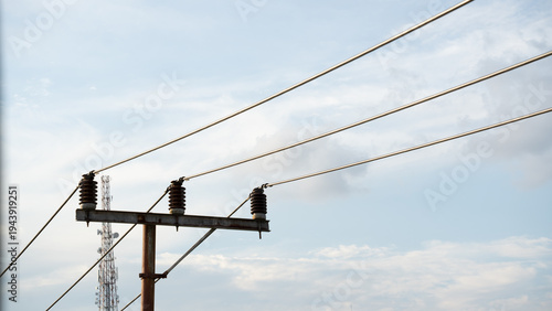A low angle shot of overhead power lines connected to ceramic insulators on a metal utility pole, with a telecommunication tower visible in the background under a bright, cloudy sky.