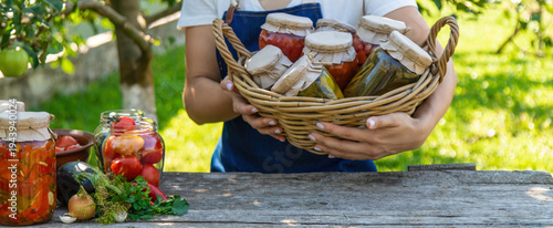 Woman holding wicker basket with jars of preserved vegetables on wooden table