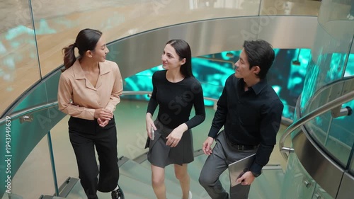 Group of young Asian business professionals smiling and talking while walking up a curved staircase in a modern office building, teamwork and corporate lifestyle concept in slow motion