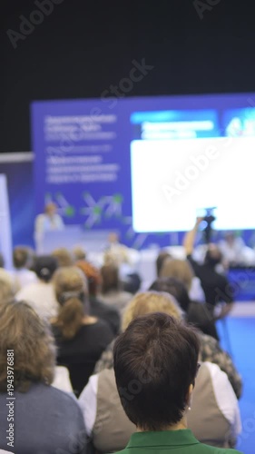 Large group of people are sitting in a room and watching a presentation. There are several people in the front row and many more in the back