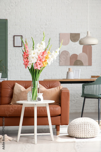 Comfortable sofa, pouf, table and vase with gladiolus flowers in interior of living room, closeup