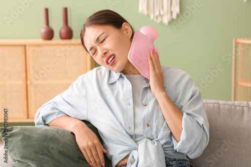 Canvas Print Young Asian woman warming her neck with hot water bottle on sofa in living room