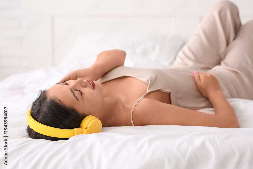 Fototapeta premium Beautiful young happy Asian woman in headphones listening music on bed at home
