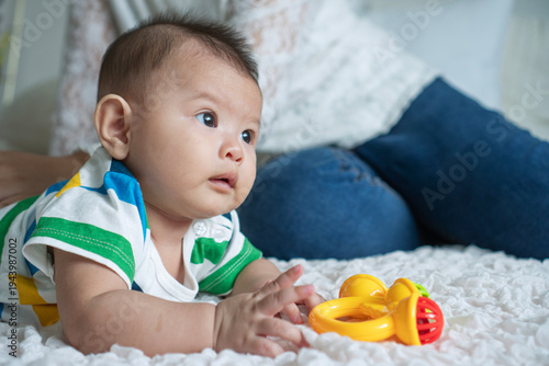 Cute Asian baby boy in striped shirt lying on tummy on white blanket looking away. Little infant playing with yellow toy rattle on bed with mother in blurred background. Parenting and care concept.