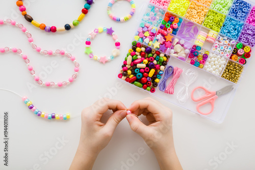 Girl hands making bracelet. Case with different colorful plastic parts. White table background. Creating different beaded jewellery. Little child handcraft. Closeup. Point of view shot. Top down view.
