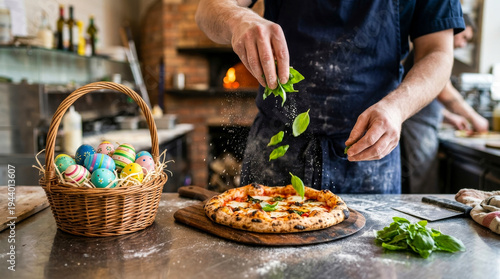 Male chef sprinkles fresh basil leaves onto a freshly baked pizza on a wooden board with a basket of colorful Easter eggs in a rustic kitchen setting