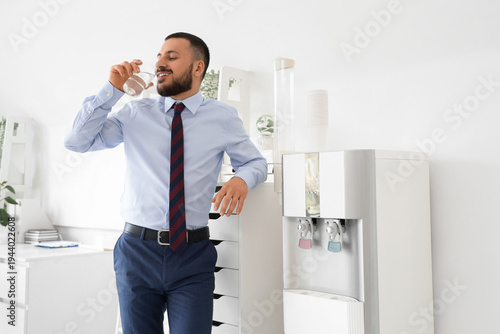 Young businessman drinking water near cooler in office