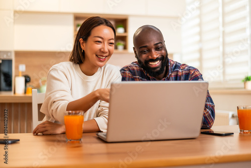 A cheerful couple sits together at a wooden table, smiling and laughing while looking at a laptop screen in a bright and cozy home kitchen.