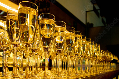 Many glasses of sparkling champagne lined up on a golden bar counter for a corporate banquet celebration. Professional luxury catering service for weddings and gala events with empty space for text.