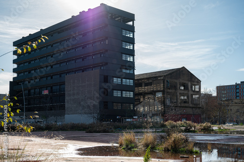 Modern black residential building standing next to a derelict industrial warehouse in a vacant urban lot with water puddles in Barcelona.