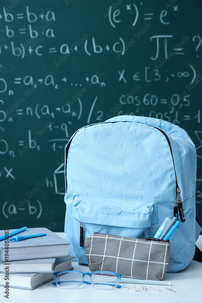 Obraz premium Blue school backpack with eyeglasses and stationery on white table near green chalkboard