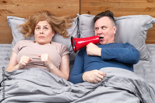 Funny couple conflict concept: annoyed man in bed shouting through red megaphone at startled woman under blanket. Humor about snoring loud wake up, relationship problems and sleepless night.