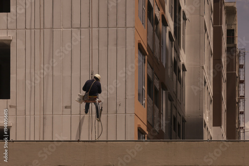 Construction worker wearing safety gear is suspended side of tall building, performing maintenance or cleaning tasks. urban setting and high rise architecture create sense of height and scale