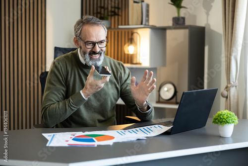 Schilderij op canvas Man using voice assistant on phone while working on laptop with business charts