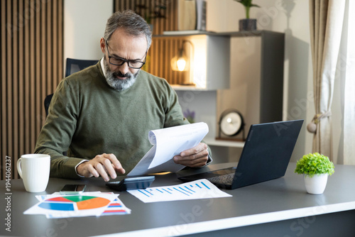 Man managing personal finances, reviewing documents, and using a calculator at his desk in a modern home office