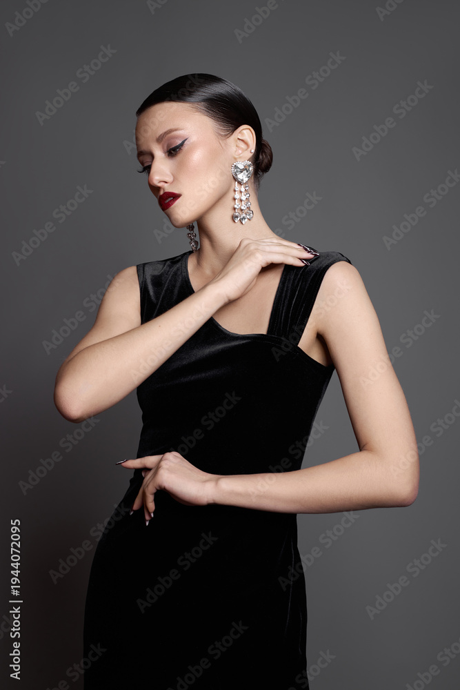 Naklejka premium Beauty fashion woman stands in studio with a black outfit. She poses with her hand near her neck. Her hair is styled back, and she wears large earrings. lighting is soft, focusing on her features