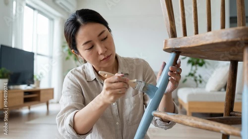 Asian woman painting a wooden chair leg with a brush in a bright living room, showcasing a modern interior with a cozy atmosphere and natural light