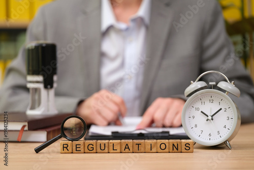 In a professional environment, a person in a suit sits at a desk, focusing on documents while surrounded by tools like a clock, magnifying glass, and books, highlighting the nature of regulatory work.