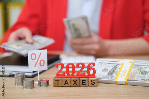 On a wooden desk, a person sorts cash while looking at a calculator, preparing for taxes due in 2026. Coins and a sign emphasize the importance of budgeting during tax season.