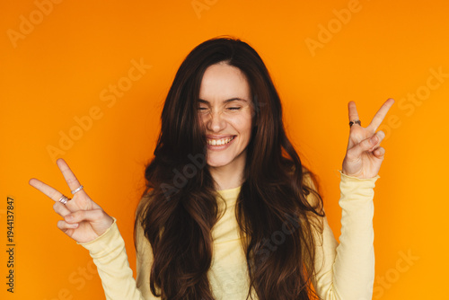 Fotografía Portrait of playful young woman showing peace sign gesture on orange background,