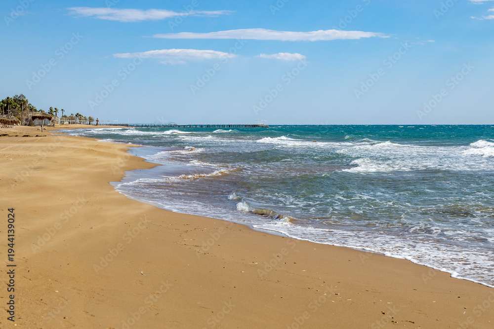 Obraz premium Wide view of a deserted sandy beach with sea waves and foam. Off-season coastal landscape with a wooden pier under a blue sky in a quiet Mediterranean resort in Sorgun, Side, Turkey.
