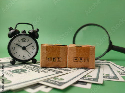 Two miniature cardboard shipping boxes placed on a fan of US dollar bills, accompanied by a small black alarm clock and a magnifying glass in the background