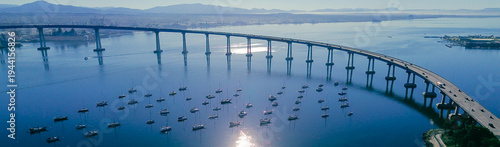 Panorama aerial curved bridge overlook frames busy Coronado Bridge above anchored boats near sandy edge of Tidelands Park and Glorietta Bay. Sunlit water patterns, palm silhouettes, shoreline, CA