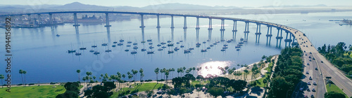 Panorama aerial park and roundabout layout with anchored bay corridor Coronado Bridge rising over San Diego Bay, moored sailboats and palm lined shoreline near Tidelands Park. Reflective water, CA
