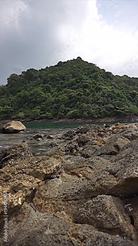 Rocky shoreline with flowing water in the foreground and a lush green hill under a cloudy sky