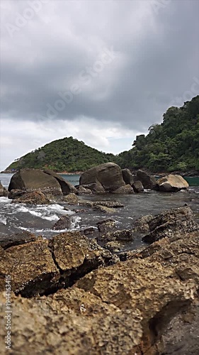 Natural landscape with rocks, water on a calm beach, and lush hills in the background.