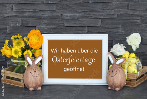Öffnungszeiten an Ostern.: Schild mit der Aufschrift  Wir haben über die Osterfeiertage geöffnet mit Osterdekoration und Blumen.