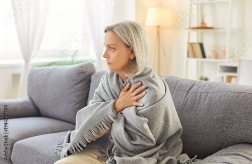 Portrait of senior gray-haired woman sitting on sofa in living room at home wrapped in cozy blanket trying to keep warm. Elderly retired person freezing in winter in chilly room with low temperature.