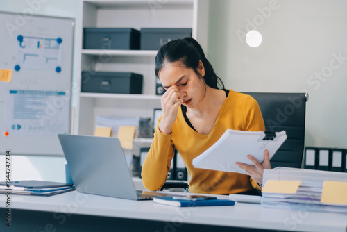 Young professional woman suffering from headache while analyzing financial charts and graphs on computer, experiencing work related stress in modern office