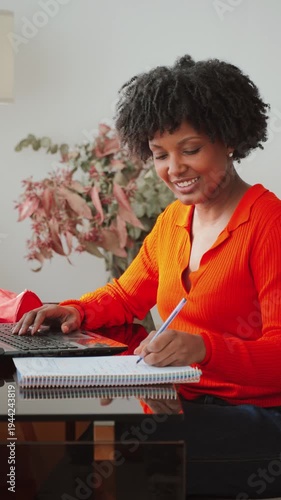 A focused woman engaged in creative writing is using a laptop while taking notes in a notebook, demonstrating productivity and concentration in her work, showcasing determination and motivation.