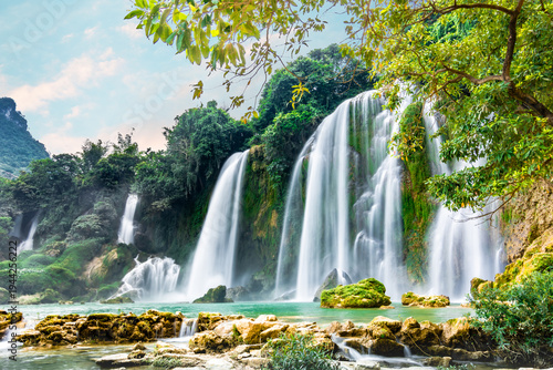 Foto Ban Gioc waterfalls tumble down stepped rock faces draped in green moss, mist rising over a calm jade pool surrounded by dense rainforest