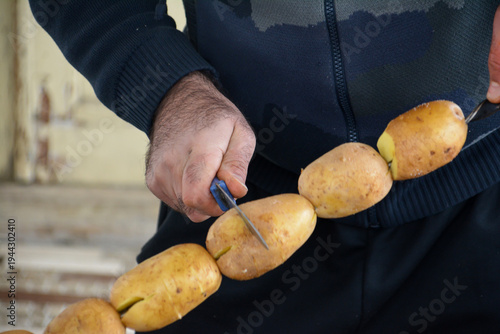 Preparing potatoes. A man in a black jacket cuts potatoes with a knife on the street. The hands of man holding a large potato in the background.