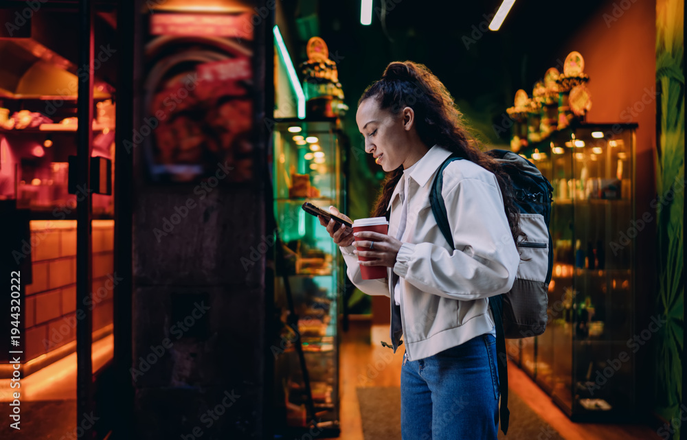 Fototapeta premium Young woman walks past colorful storefronts, holding smartphone and coffee, immersed in digital routine while navigating urban evening filled with artificial light.