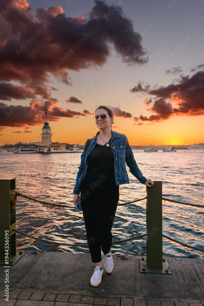 Fototapeta premium Mujer feliz al atardecer con la icónica Torre de la Doncella (Kız Kulesi) de fondo en Estambul. Cielo dramático y nubes coloridas sobre el Bósforo durante la hora dorada.