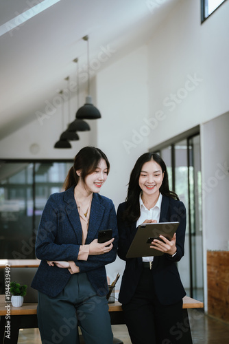 Two professional businesswomen discussing work in a modern office, one using a smartphone while the other holds a tablet, collaborating in a bright, contemporary workspace.