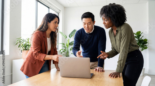Three diverse coworkers brainstorming ideas while standing around a laptop with back side visible, modern workspace with plants and natural light.