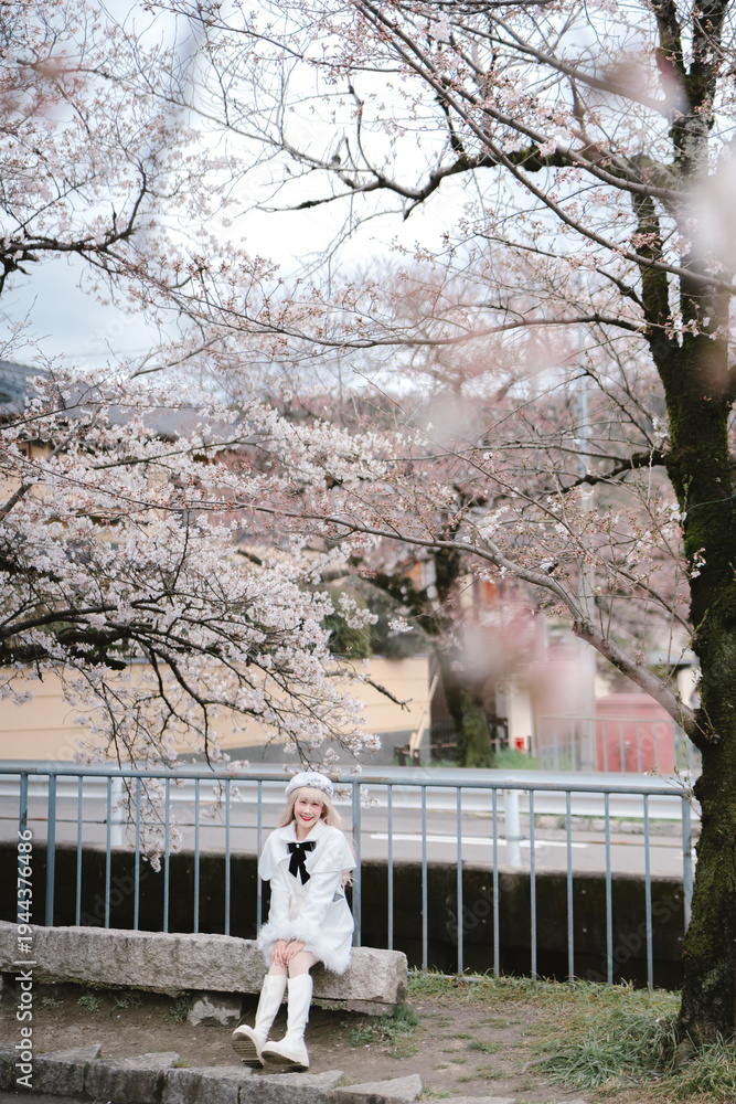 Fototapeta premium Young woman sitting on stone bench under blooming cherry blossom tree in spring park, wearing white dress and boots, enjoying peaceful outdoor moment with soft natural light